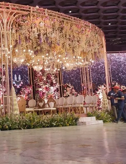 A wide view of the floral reception stage during setup, revealing the scale of the golden structure and the army of florists required to bring this vision to life.