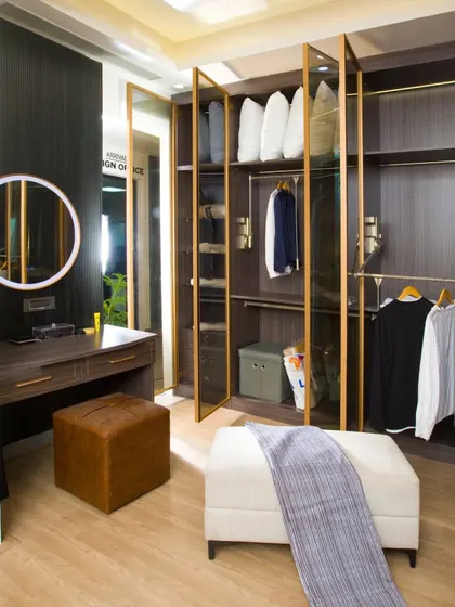 A luxurious walk-in closet and dressing area. The design features glass-front wardrobes, a dedicated vanity with a lit mirror, and an ottoman for seating.