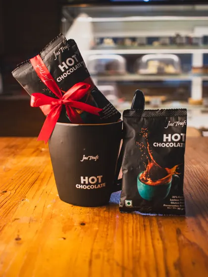 Our Hot Chocolate Mix sachets sit in a black mug with a red ribbon, beside another pack on the café counter.