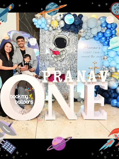 A happy family photo from a space-themed first birthday. The backdrop includes a silver sequin wall, blue balloon arrangements, and an astronaut prop.