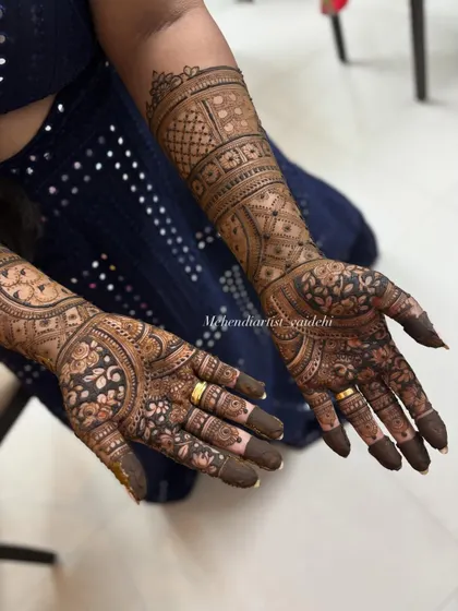 A bridal mehendi design that extends up the forearms. The right hand features floral and paisley patterns, while the left arm showcases geometric grids and checks for a unique, asymmetrical look.