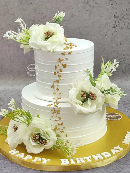 An elegant two-tiered cake for an 80th birthday. The clean white design is adorned with beautiful white flowers and a sprinkle of gold flakes.