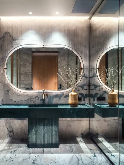 The story of elegance continues in the bathrooms at Palais Royale, where clean lines and precision detailing are paramount. This bathroom features dual vanities in a striking fluted green stone, set against a backdrop of book-matched marble and backlit oval mirrors.