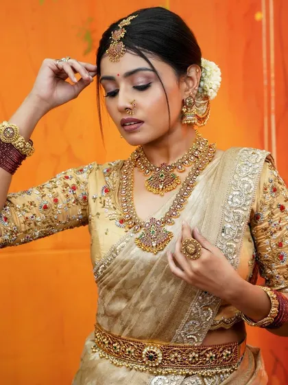 A candid moment capturing the grace of a bride adjusting her jewelry. This cream and gold saree is a testament to timeless bridal fashion, perfect for creating lasting memories.