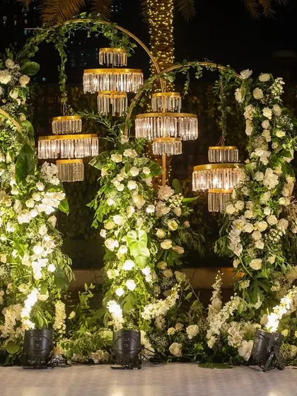 A romantic and elegant photo backdrop for Yashvi and Siddharth's celebration. The setup features arches of white flowers and greenery, accented with modern crystal light fixtures.