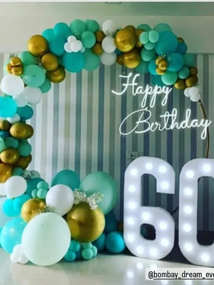 Celebrating a 60th birthday with a stylish ring backdrop. The teal, white, and gold balloon garland is paired with a "Happy Birthday" neon sign and large marquee numbers for a memorable setup.