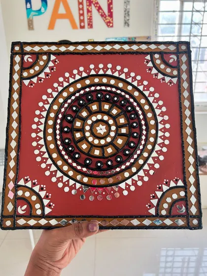 This square Lippan art piece features a traditional design in earthy red and brown tones, with intricate mirror work.