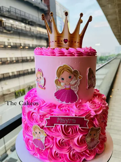 A two-tier princess cake perfect for a royal celebration. The bottom tier is decorated with pink buttercream rosettes, while the top tier is smooth pink, all crowned with a princess tiara.
