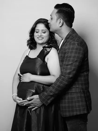 A classic black and white couple's portrait. The father-to-be, wearing a plaid blazer, embraces his partner, who is in a dark gown, creating a timeless and elegant image.