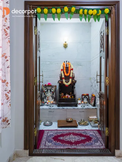 A traditional pooja room with intricately carved wooden doors. Inside, a marble platform and simple wall sconce create a serene and timeless space for daily worship.