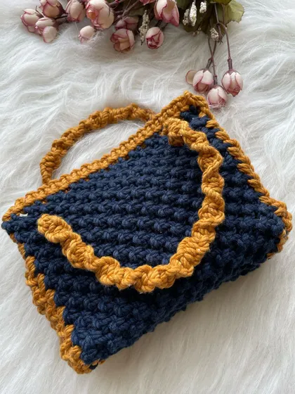 This handle bag is made with a dense, textured knot in a deep navy blue, accented with a ruffled tan border and a twisted handle. It's spacious and holds its shape well.