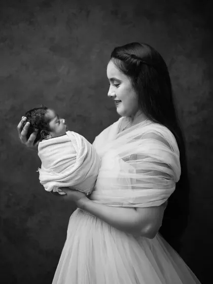 An elegant black and white portrait of a mother in a flowing gown holding her swaddled newborn. The dark background and soft lighting create a timeless, fine-art feel for this newborn photography session.
