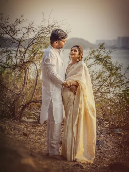 An expecting couple, dressed in elegant white traditional wear, looks at each other lovingly by the water. The soft, hazy light gives the image a timeless and romantic feel.
