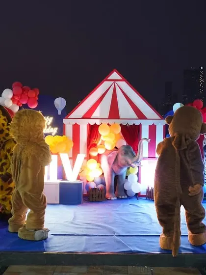 A wide shot of a circus performance at night, featuring multiple mascots and performers on a stage with a classic red-and-white tent backdrop.