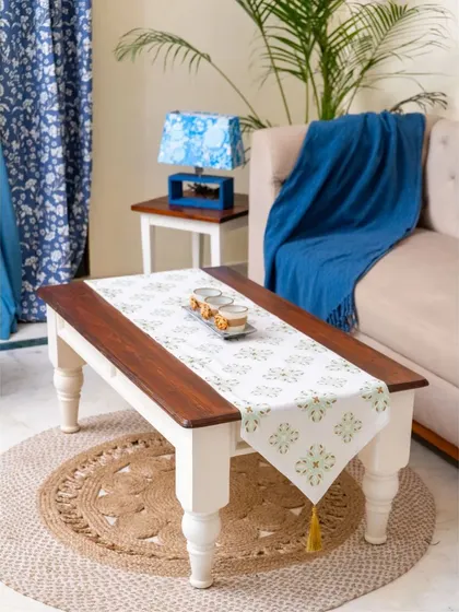 A farmhouse-inspired coffee table featuring a rich wooden top and distressed white turned legs. Placed on a natural jute rug, it adds a touch of rustic charm and elegance to the living space.