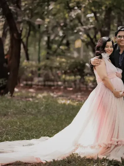 An enchanting outdoor portrait of an expecting couple. The mom-to-be's long, flowing pink gown trails behind her as they embrace amidst the greenery of a park.