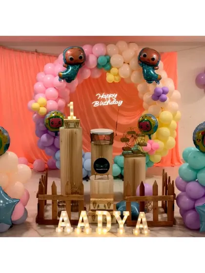 A pastel Cocomelon theme for a first birthday, featuring a circular balloon arch, character foil balloons, and a picket fence. The setup is personalized with the name 'Aadya' in marquee letters.