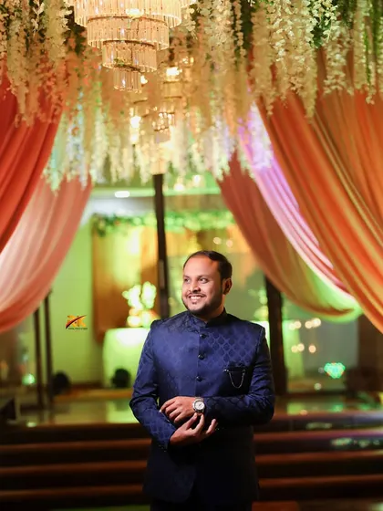 The groom stands under a grand chandelier and floral ceiling decoration at the reception venue. The ambient lighting creates a warm and celebratory mood for this solo portrait.