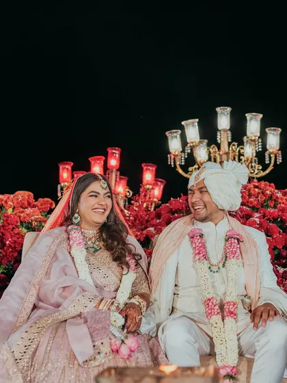 The couple shares a laugh on their wedding stage, surrounded by a lush arrangement of red flowers and the warm glow of candelabras.