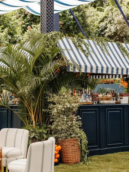 A custom-built bar designed for an outdoor birthday party. The navy blue paneling and striped awning are surrounded by lush palm fronds and potted plants, seamlessly blending the service area into the garden theme.