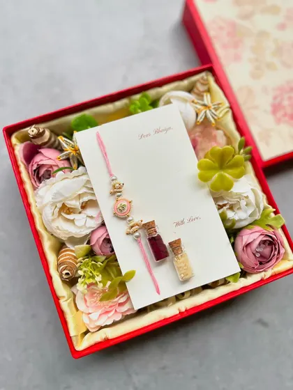 The inside of one of our Rakhi gift boxes, featuring a beautiful Rakhi and Lumba set on a personalized card, surrounded by a bed of artificial flowers and traditional Roli Chawal vials.