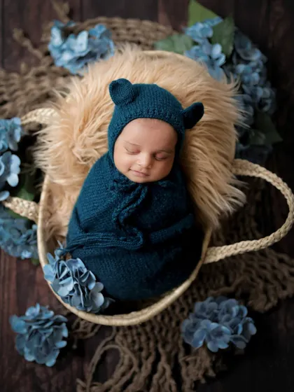 A little bear cub sound asleep. This newborn is wrapped snugly in a basket, surrounded by blue flowers, creating a dreamy and adorable portrait.