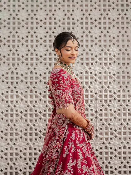 A serene portrait of the bride in her royal red lehenga, standing against an intricately designed wall. Her gentle smile and elegant posture reflect the timeless grace of a royal bride.