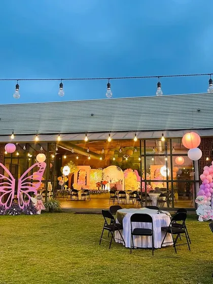 An outdoor view of the enchanted garden party, with a large pink butterfly prop and string lights creating a magical ambiance as evening approaches.