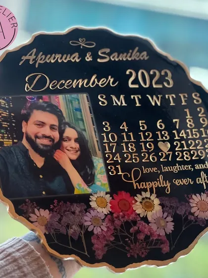 A modern and chic black resin calendar frame. This piece highlights a December 2023 wedding with the couple's names, photo, and a beautiful quote, surrounded by preserved wildflowers.