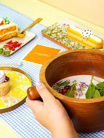 A lifestyle shot showing hands holding a wooden serving bowl, with a table set with colorful plates and pastries in the background. This adds a human element to the scene.