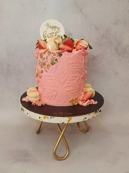 A tall cake with a textured pink leaf pattern, decorated with fresh strawberries, macarons, and a touch of gold leaf.