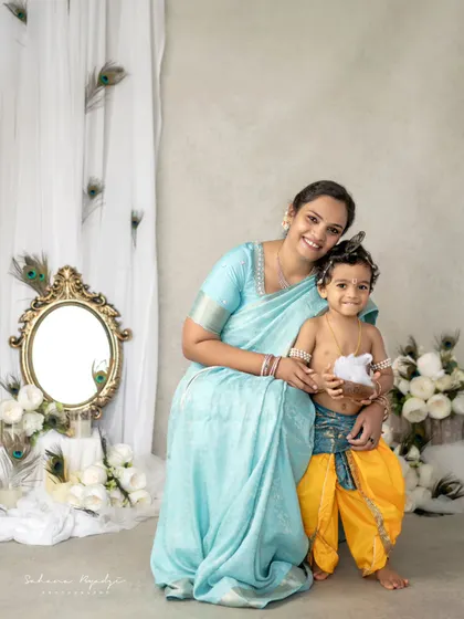A beautiful portrait of a mother and her son in a Krishna-themed setup. The peacock feather decorations and elegant mirror add a touch of royalty to this special photo.