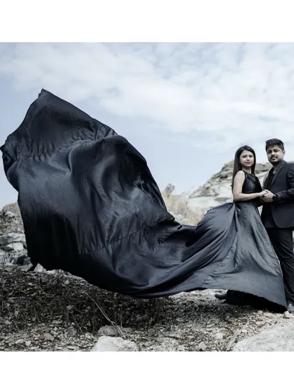 A powerful wide shot of a couple in a rugged, rocky setting, with the black fabric of her trail gown caught in the wind, creating a cinematic effect.