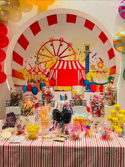 A candy bar at a carnival-themed party. The table is laden with jars of colorful candies and treats, set against a backdrop featuring a Ferris wheel and circus tent.