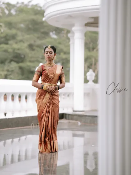 A full-length portrait of the bride in her beautiful copper-toned Muhurtham saree. The classic white balcony provides a clean and elegant backdrop for this graceful shot.