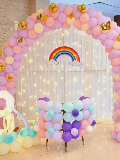 A beautiful pastel rainbow balloon arch with a unicorn theme. The fairy lights behind the curtain add a magical twinkle to the entire setup.