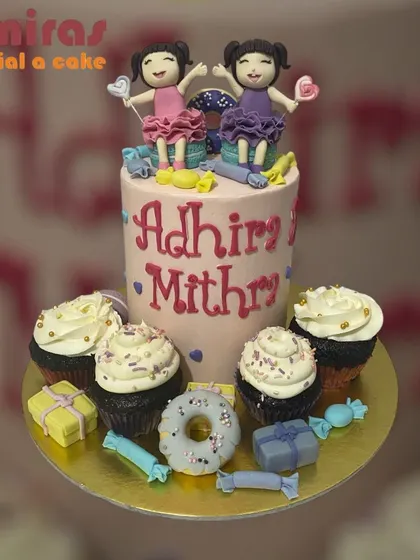 A fun cake and cupcake set for twins, Adhira and Mithra. The main cake is topped with figurines of the two girls, surrounded by matching cupcakes and sweet treats.