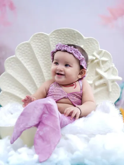 A happy baby girl in a lavender mermaid costume, smiling brightly from her seashell throne.