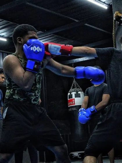 A senior boxer spars with a junior, showing the ropes. The whole team watches, learning from every exchange in the ring.