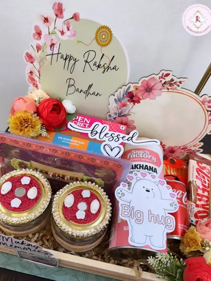 A beautiful Rakhi hamper designed to make your sibling feel blessed. This tray includes decorated jars, snacks, a drink, and a custom "Happy Raksha Bandhan" sign, all arranged with floral accents.