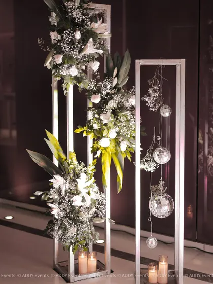 We use modern structures like these white metal frames to create unique floral installations. Adorned with white lilies, baby's breath, and hanging disco balls, they serve as artistic decor pieces for a contemporary event.