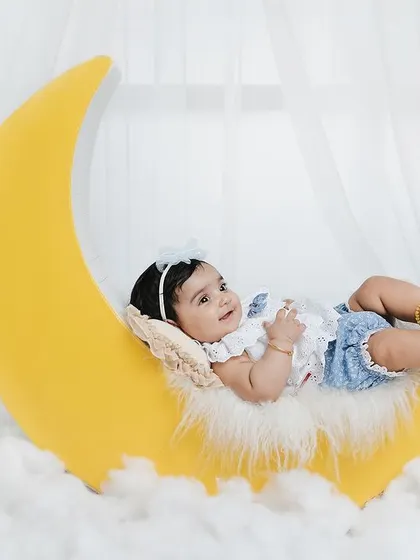 A whimsical portrait of a baby resting on a large yellow moon prop, surrounded by fluffy white clouds. This dreamy studio setup is perfect for capturing magical baby photos.