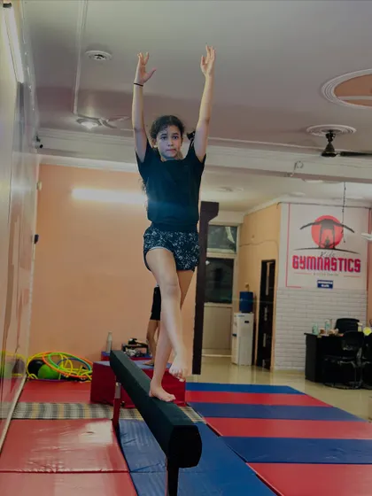 A student demonstrates perfect form and balance on the beam. Her arms are raised high as she prepares for her next move, showing great concentration.