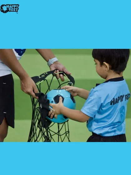 Teamwork makes the dream work, even when it comes to packing up! A child helps a coach put a football into the bag, learning about responsibility and helping out after a fun session.