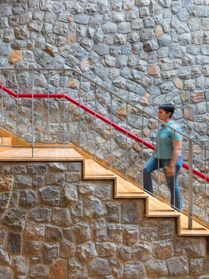A staircase with wooden treads and a bright red handrail runs alongside a massive wall of locally sourced random rubble stone, a key material in the project.