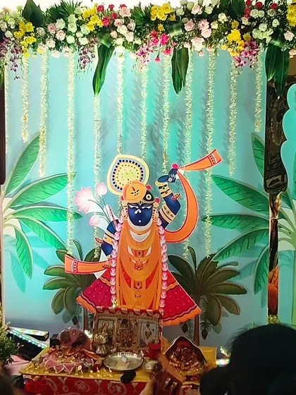 A beautiful backdrop for a home bhajan program dedicated to Shrinathji. The decor features a custom printed backdrop with banana tree illustrations and a floral arrangement on top.