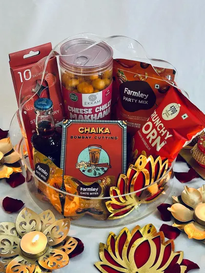 Embrace the auspicious color red with this vibrant Diwali hamper. Housed in a clear acrylic basket, it's filled with red-packaged snacks, drinks, and festive lotus-themed decor.