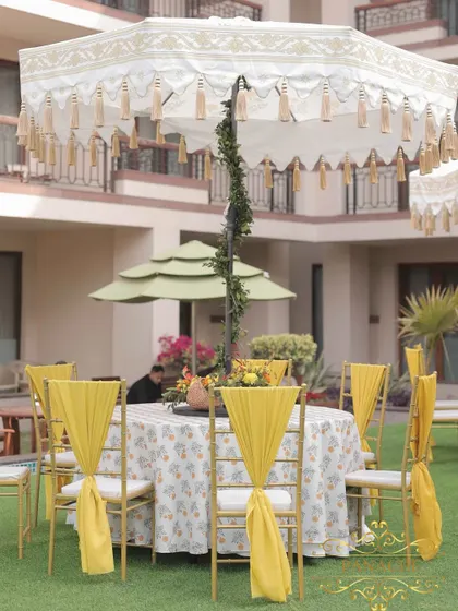 Guest seating for the Haldi, with printed tablecloths, yellow chair drapes, and large decorative umbrellas providing shade and style.