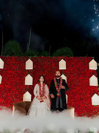 The couple stands amidst a cloud of low fog on their reception stage, with a backdrop of vibrant red flowers and the glow of cold pyrotechnics.