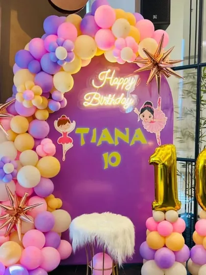 A ballerina-themed 10th birthday party. This setup includes a purple backdrop, ballerina cutouts, a "Happy Birthday" neon sign, and a pastel balloon arch.